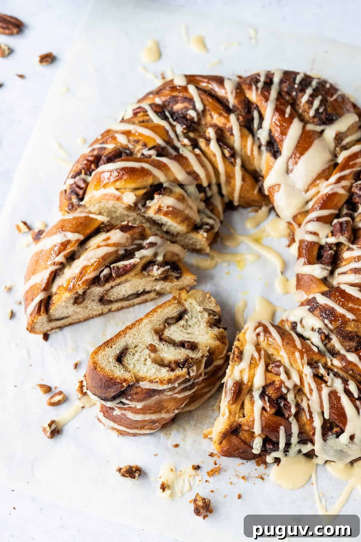 Maple pecan bread wreath – this impressive vegan maple pecan couronne is a real showstopper which would be perfect for an indulgent breakfast or brunch. #veganbaking #eggless #dairyfree #bread #veganbreakfast #veganbrunch Close up of sliced maple pecan bread wreath drizzled with maple glaze on a sheet of baking parchment.