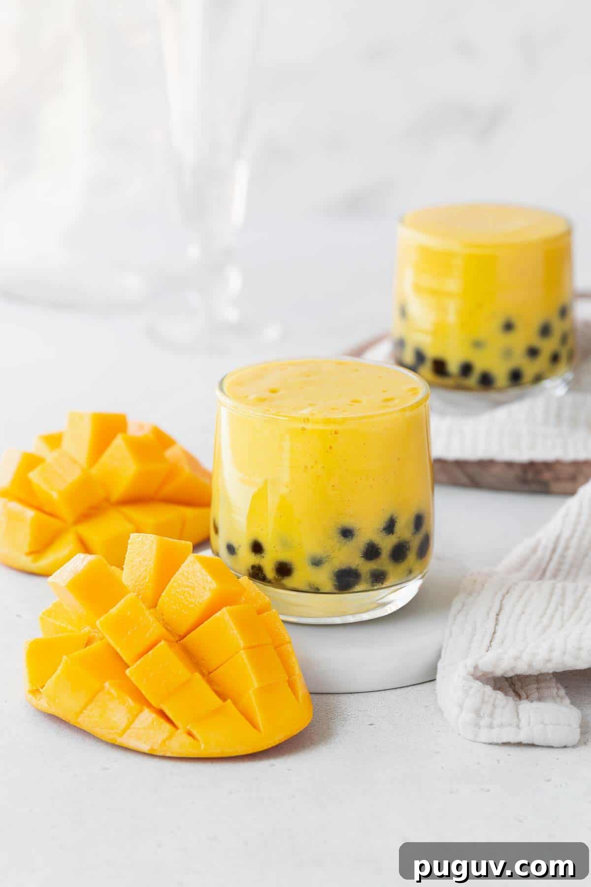 Two glasses of mango boba tea on a white counter next to a cubed up mango.
