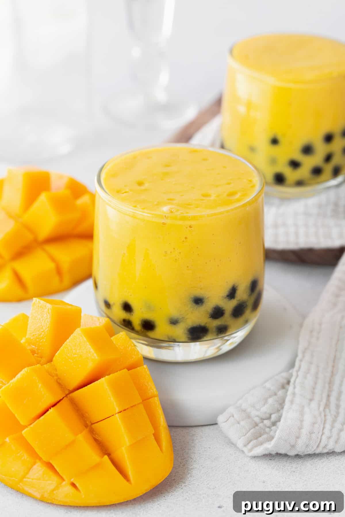 A closeup of a small glass of mango boba tea next to a cubed mango and towel.