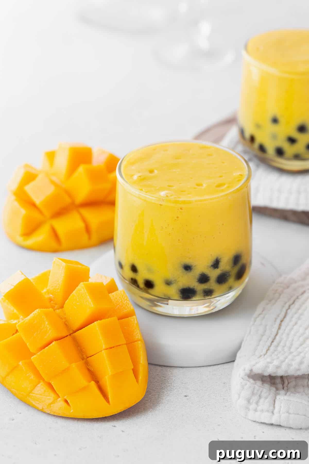 A closeup shot of a small glass of mango boba tea on a white coaster on the counter next to a sliced up mango.