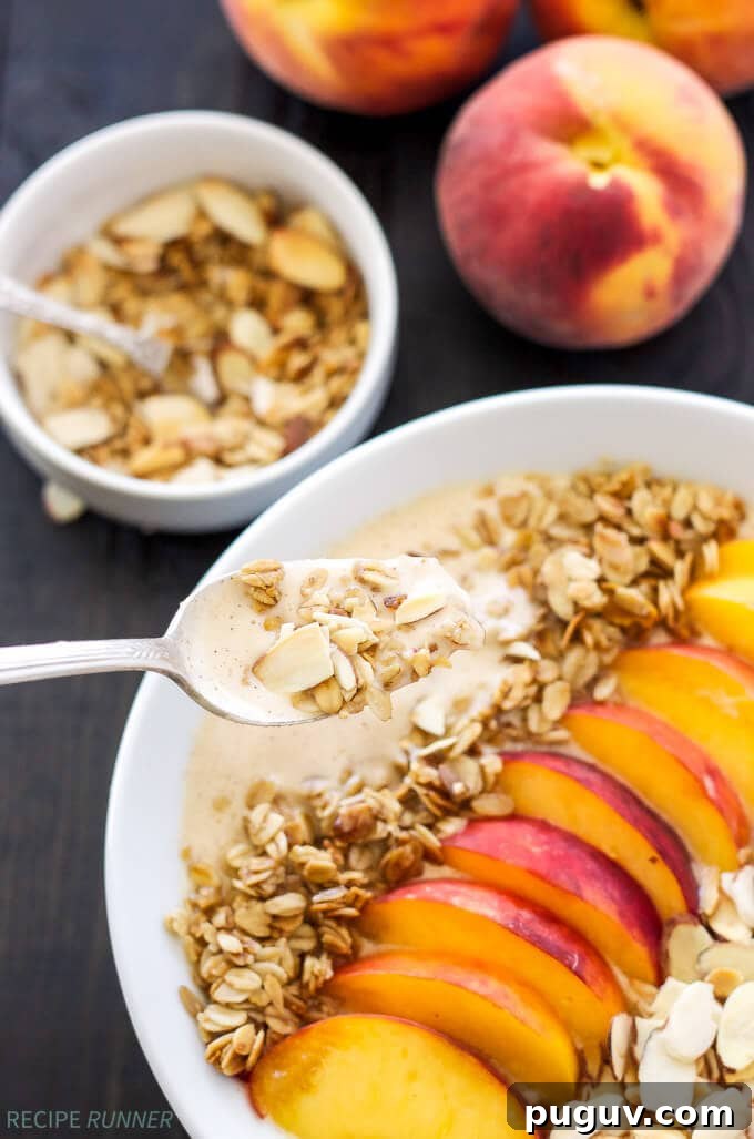 Creamy Peach Smoothie Bowl Recipe for Summer Breakfast 3 A spoon scooping into a bowl of peach smoothie with toppings like fresh sliced peaches and sliced almonds.