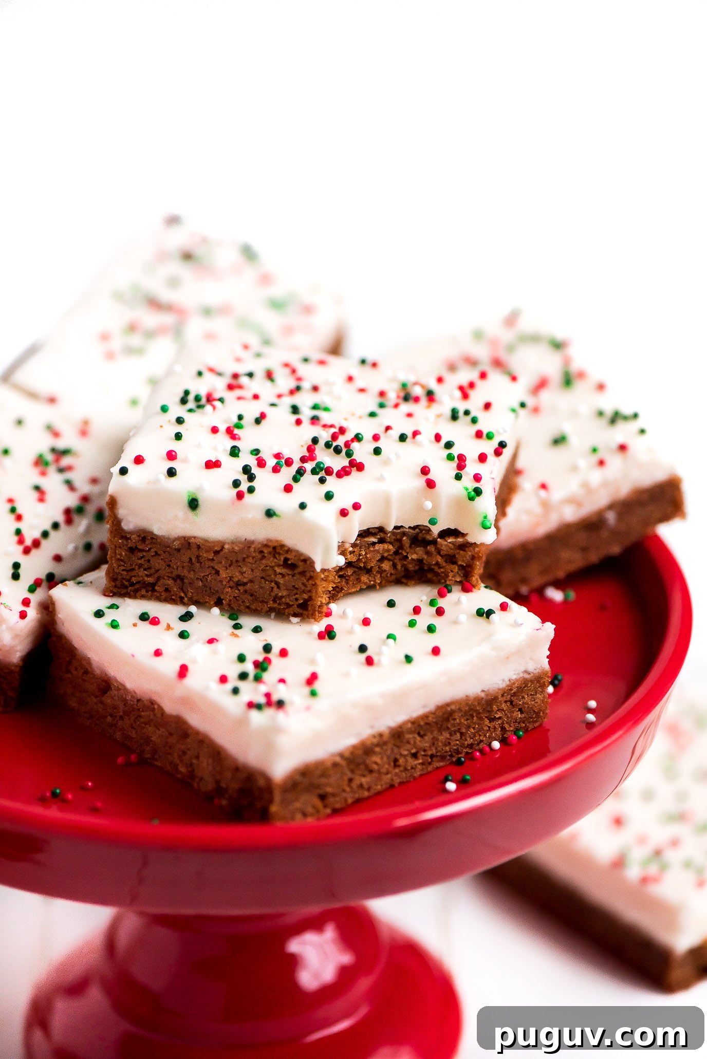These soft and chewy Gingerbread Sugar Cookie Bars topped with cream cheese frosting are so quick and easy to make. They're the perfect festive treat to share this season. Gingerbread Sugar Cookie Bars on a red cake stand with a bite taken out of one.
