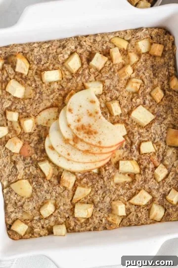 Baked apple oatmeal topped with apple slices in a square white baking dish with apples and syrup on the side.