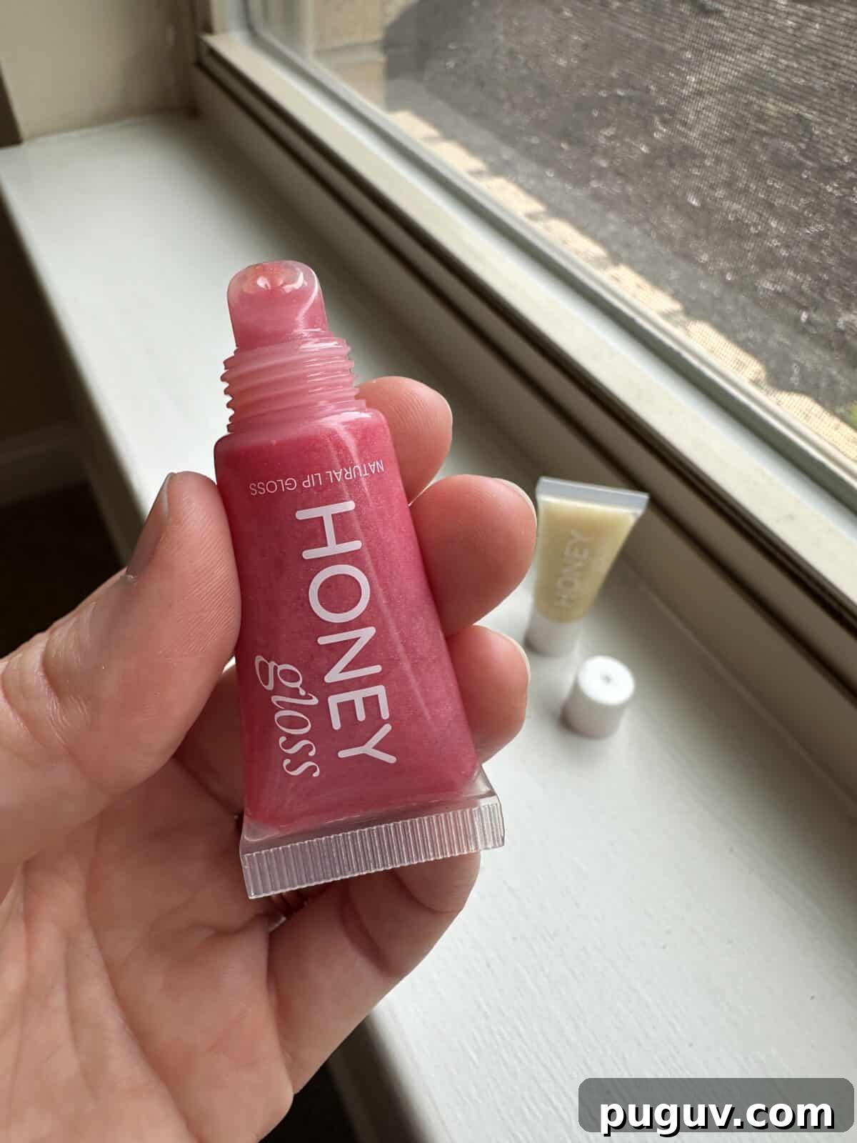 Clean Makeup for Tweens: 8 Safe Brands for Kids and Teens 6 A person's hand holds a pink squeeze tube of Honey Gloss lip gloss near a sunlit window sill, with a second tube of lip gloss visible and out of focus in the background.