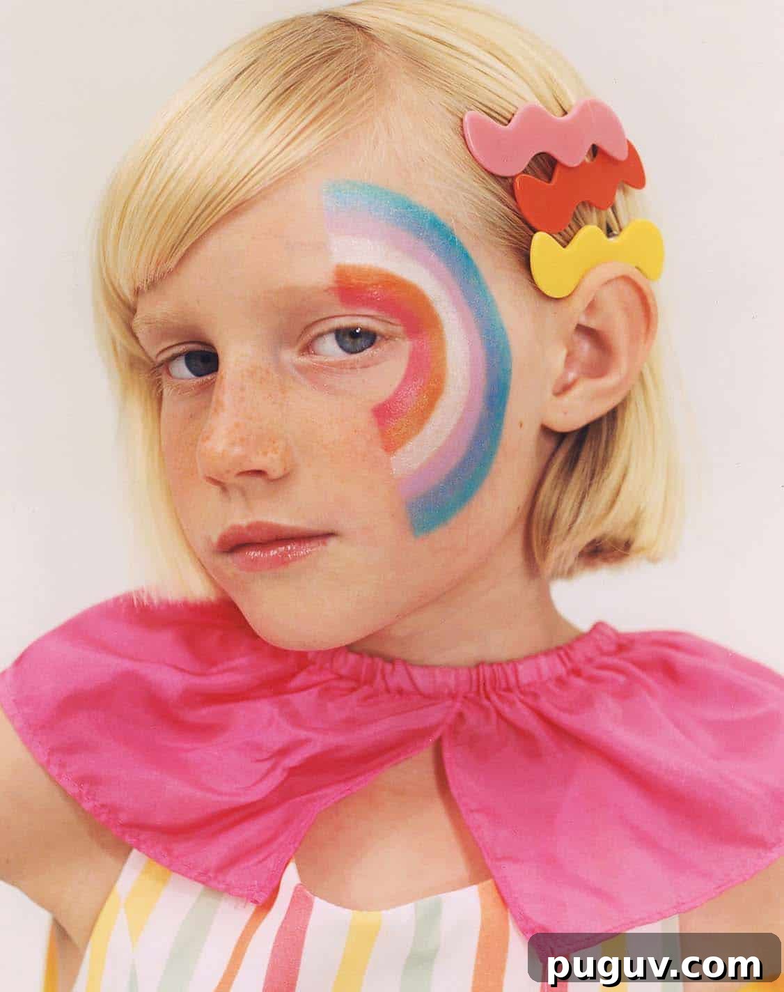Clean Makeup for Tweens: 8 Safe Brands for Kids and Teens 9 A blonde child with a serious expression and a pink ruffled collar has a colorful rainbow face painting arching from the eye across the cheek, and two multicolored hair clips are styled near their ear.