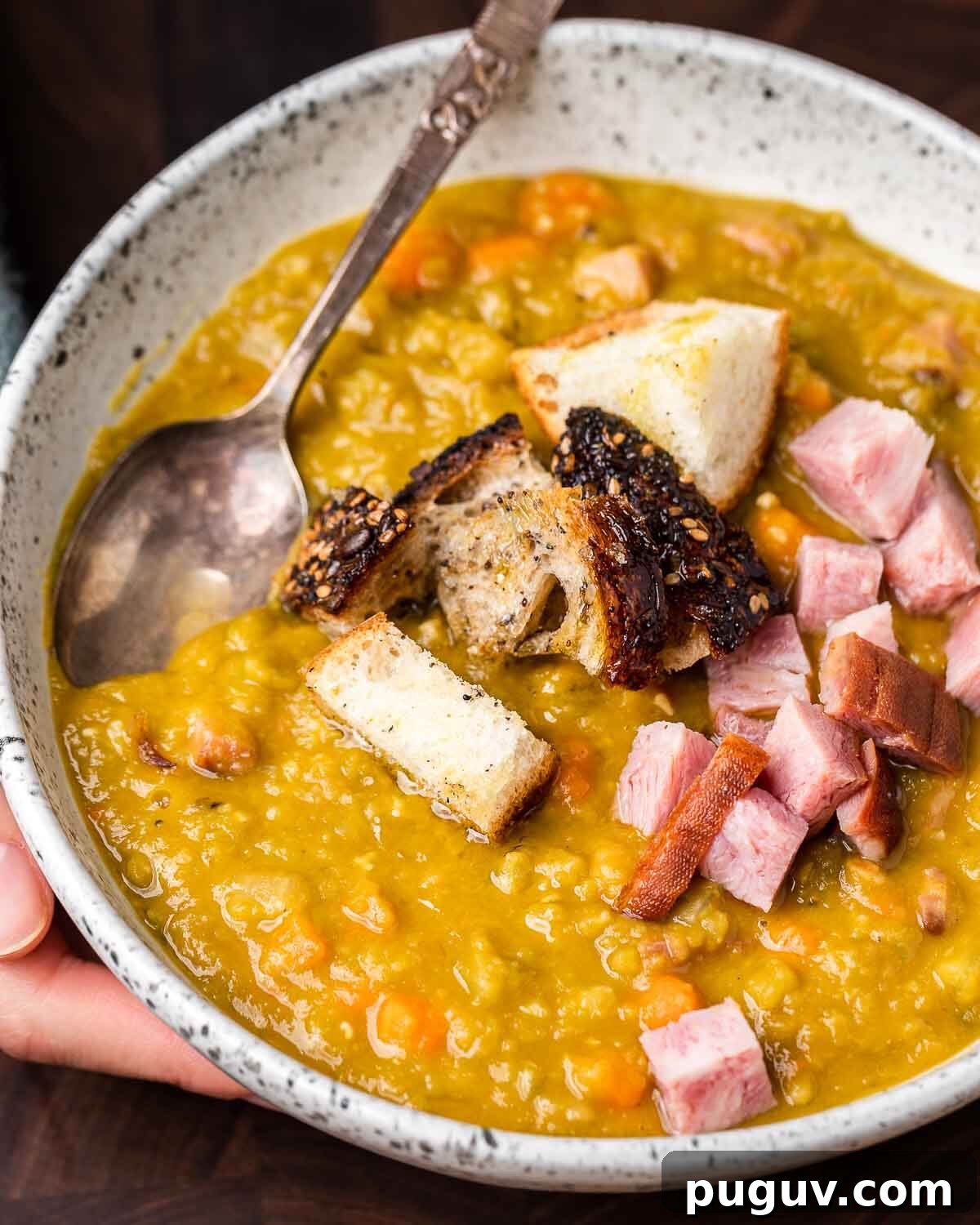 Split Pea Soup with Ham Recipe — Hearty Comfort Soup 2 Hands holding white bowl of split pea soup with ham and croutons.