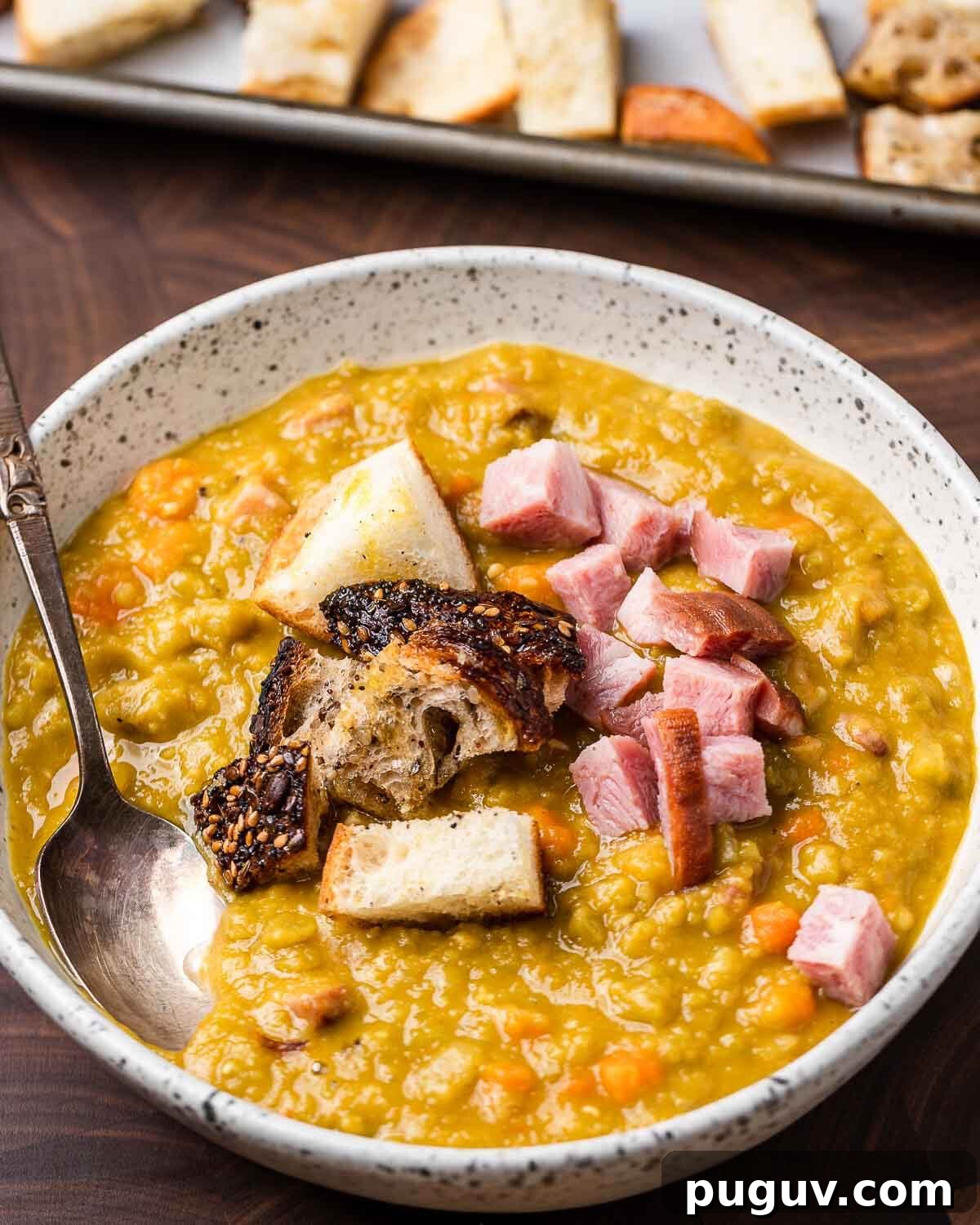 Split Pea Soup with Ham Recipe — Hearty Comfort Soup 7 White bowl of split pea soup and tray of croutons in background.