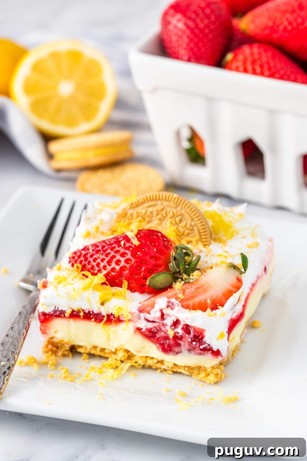 A slice of strawberry lemon cheesecake bars, with layers of strawberries, cream, and biscuit on top, garnished with lemon zest, is served on a white plate with a fork. Fresh strawberries and a lemon are visible in the background.
