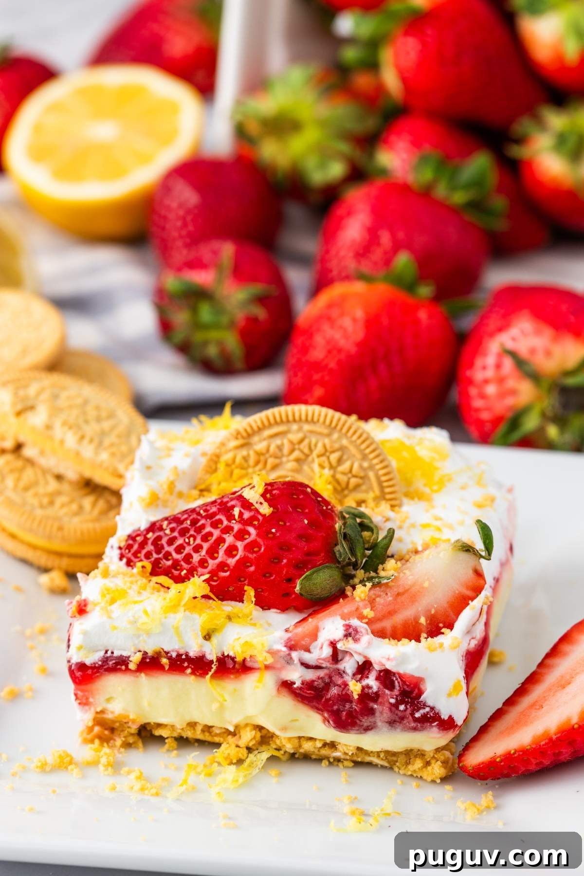 A slice of no bake strawberry lemon cheesecake bars topped with fresh strawberries, lemon zest, and a golden cookie, surrounded by strawberries, lemon slices, and cookies on a white plate.
