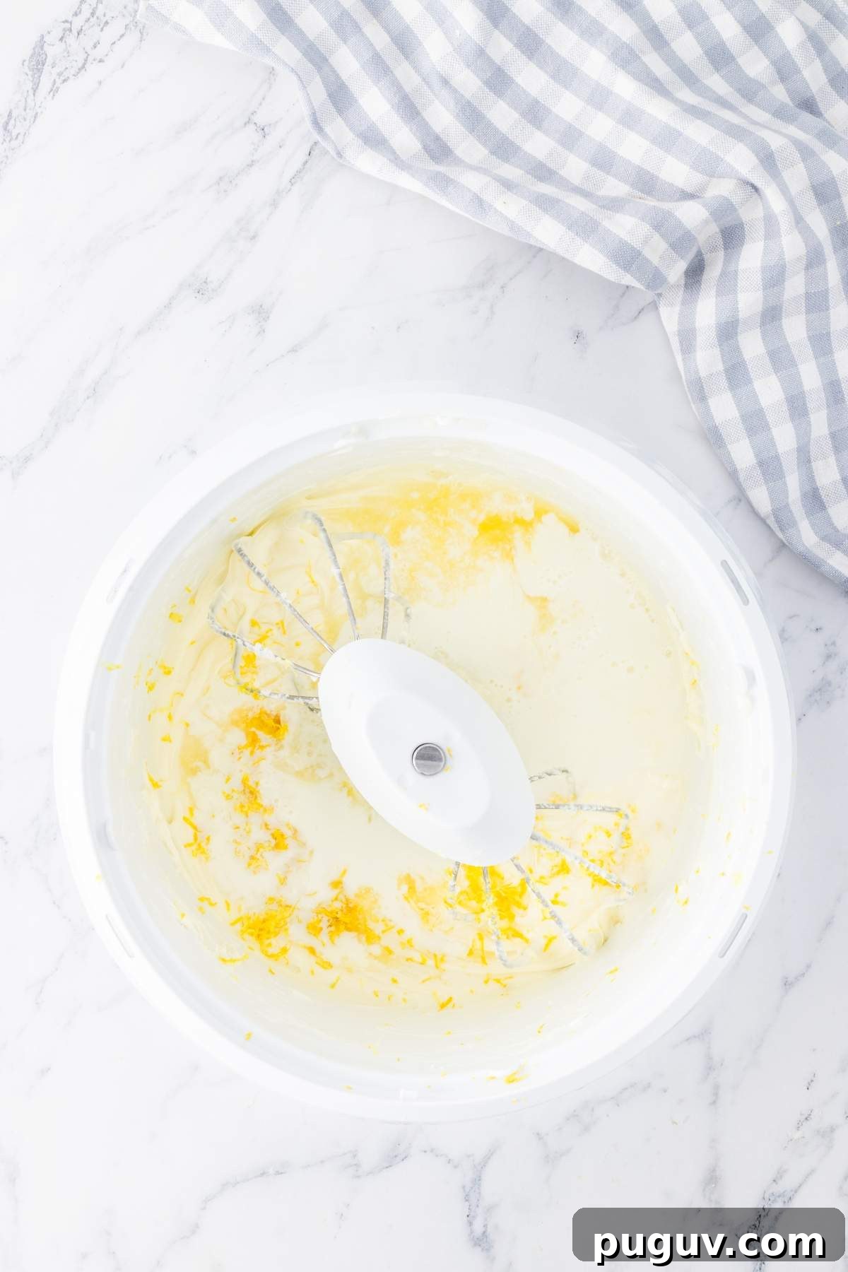 A mixing bowl with a mixer attachment sits on a marble countertop. The bowl contains partially mixed ingredients, hinting at a delicious strawberry lemon cheesecake lasagna in progress. A gingham cloth lies beside the bowl, adding to the cozy kitchen scene.