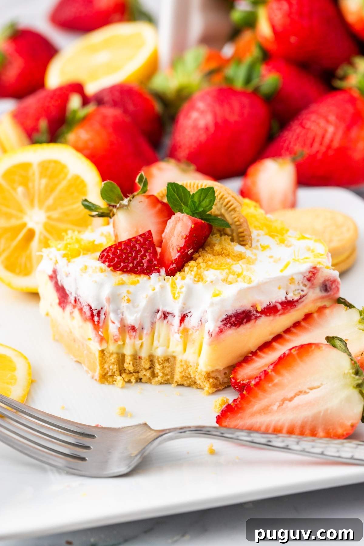 A no bake layered strawberry lemon cheesecake lasagna with a crumbly base, creamy middle, and topped with strawberries and cream. Surrounding are whole strawberries, lemon slices, and a fork on the side.