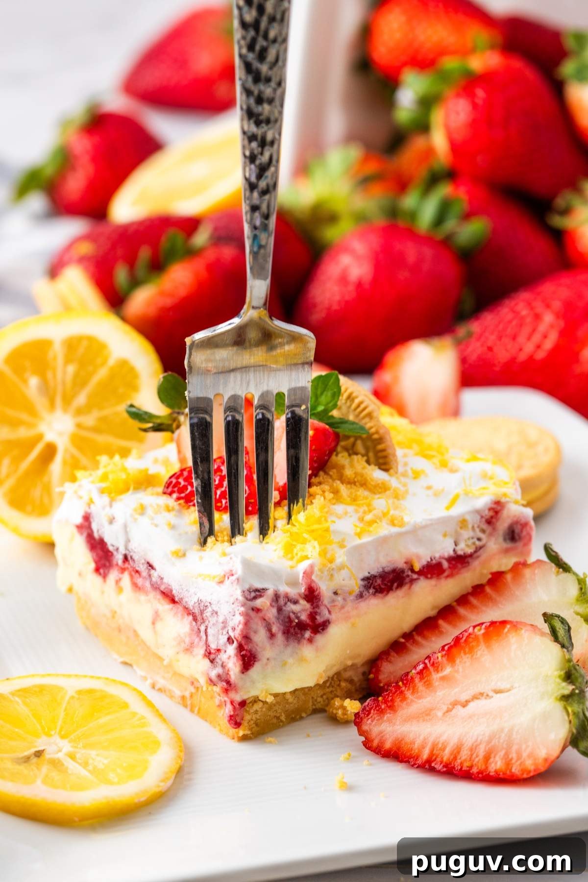 A fork is inserted into a slice of strawberry dessert lasagna with a crumbly base, cream, fruit filling, and whipped top. Sliced strawberries, lemon wedges, and whole strawberries decorate the plate.