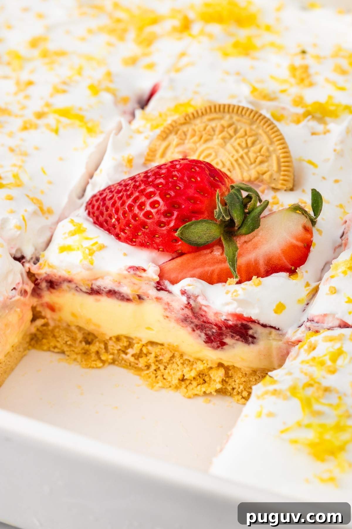 A pan with slices of a layered no bake strawberry lemon dessert topped with whipped cream, lemon zest, a golden cookie, and a fresh strawberry. One piece from the bottom left corner is missing.