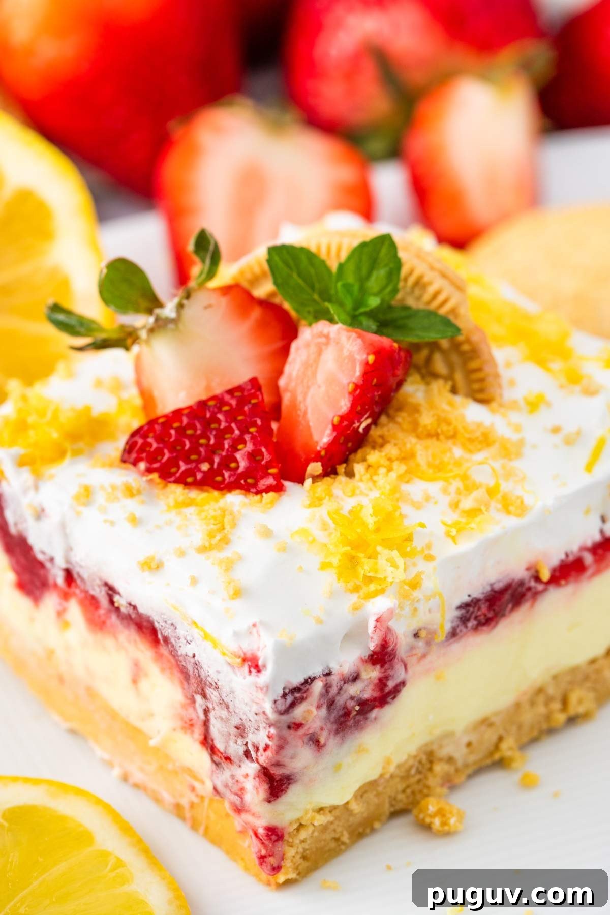 A square of layered no bake strawberry dessert with a crumbly base, creamy middle, and strawberry topping, garnished with fresh strawberries and a lemon slice.
