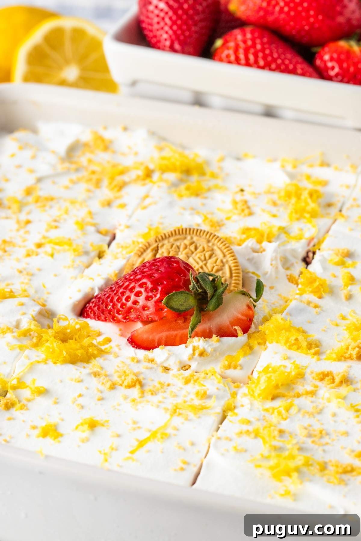 A white rectangular strawberry lemon cheesecake lasagna topped with a layer of whipped cream, lemon zest, and a single sliced strawberry with a yellow cookie. A bowl of whole strawberries is in the background.