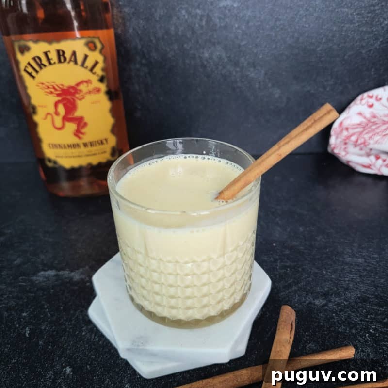Christmas Cocktail Recipes Everyone Will Love 23 Fireball Eggnog in a crystal glass with cinnamon stick garnish in front of a bottle of Fireball Cinnamon Whiskey