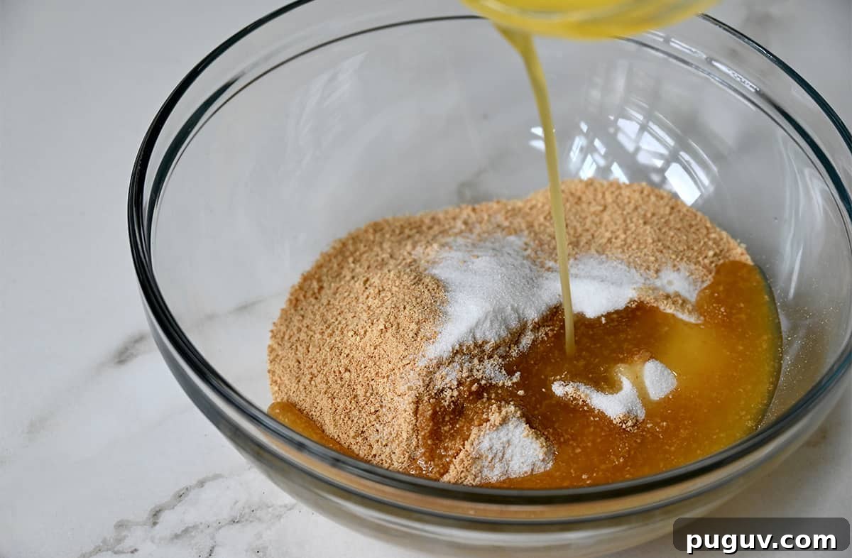 No-Bake Cheesecake Recipe: Creamy No-Bake Cheesecake Slice 2 Melted butter being poured atop crushed graham crackers and sugar in a clear bowl.