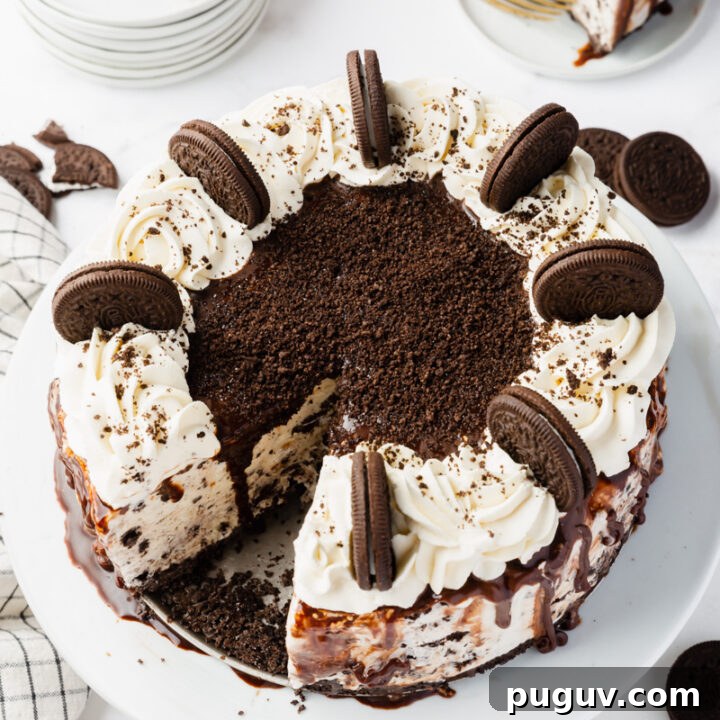 a large homemade ice cream cake topped with whipped cream and whole oreo cookies, stood up around the edge. The cake is on a plate. Behind it is a small plate with a slice of oreo ice crema cake, and a stack of small plates and two gold forks.