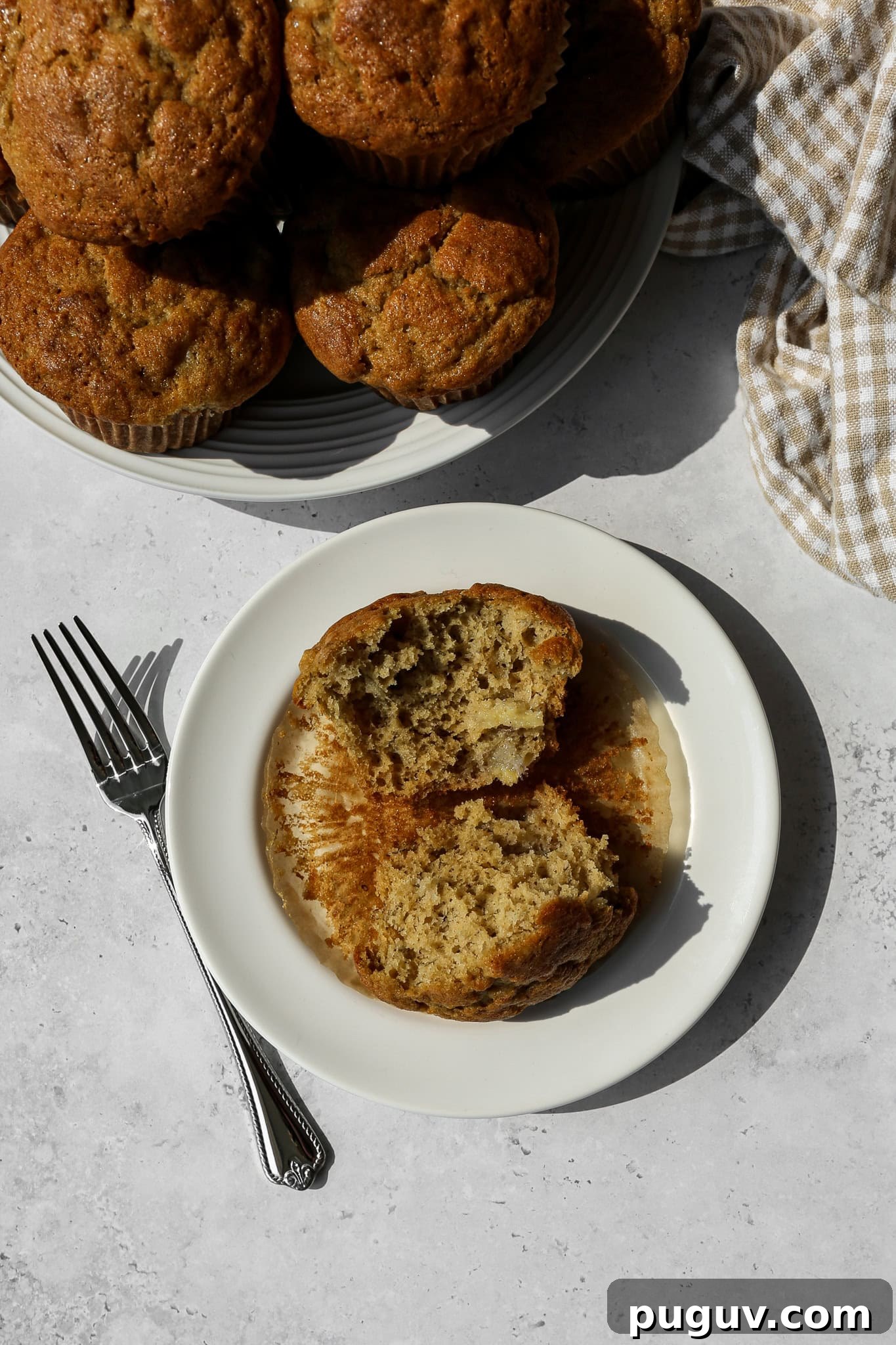 a gluten free banana bread muffin torn in half on a white plate next to a silver fork and a plate of muffins