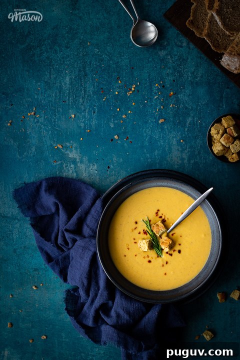 Flat lay view of spicy roast butternut squash soup with a spoon in a blue bowl set on a blue plate with a dark blue linen napkin on the side. Set on a teal backdrop, there's also a board with sliced bread, a pot with croutons and two soup spoons in the background.