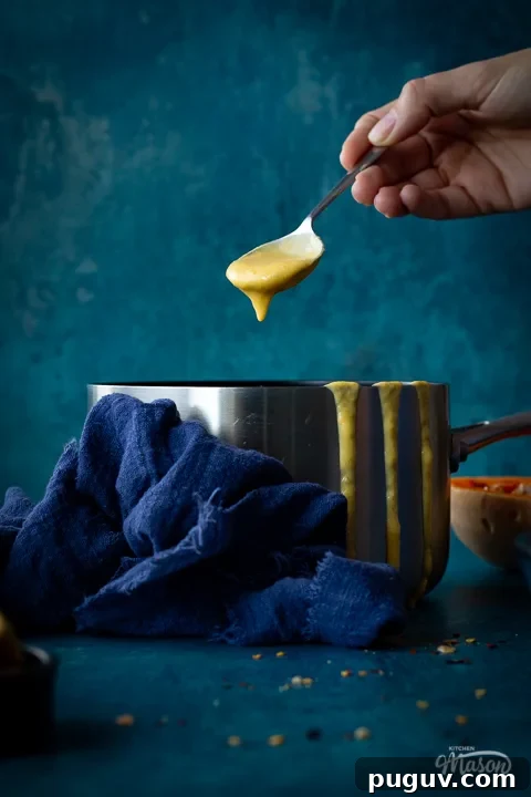 Someone reaching in a saucepan of butternut squash soup with a metal soup spoon set against a teal backdrop. There are drips of soup down the side of the pan, half a butternut squash, a pot of croutons and a blue linen napkin in the backgtround.