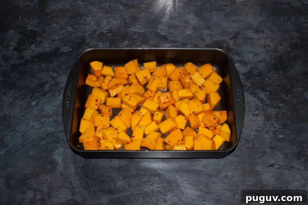 Butternut squash cubes in a roasting pan mixed with oil, chilli flakes and seasoning. Set on a kitchen worktop.