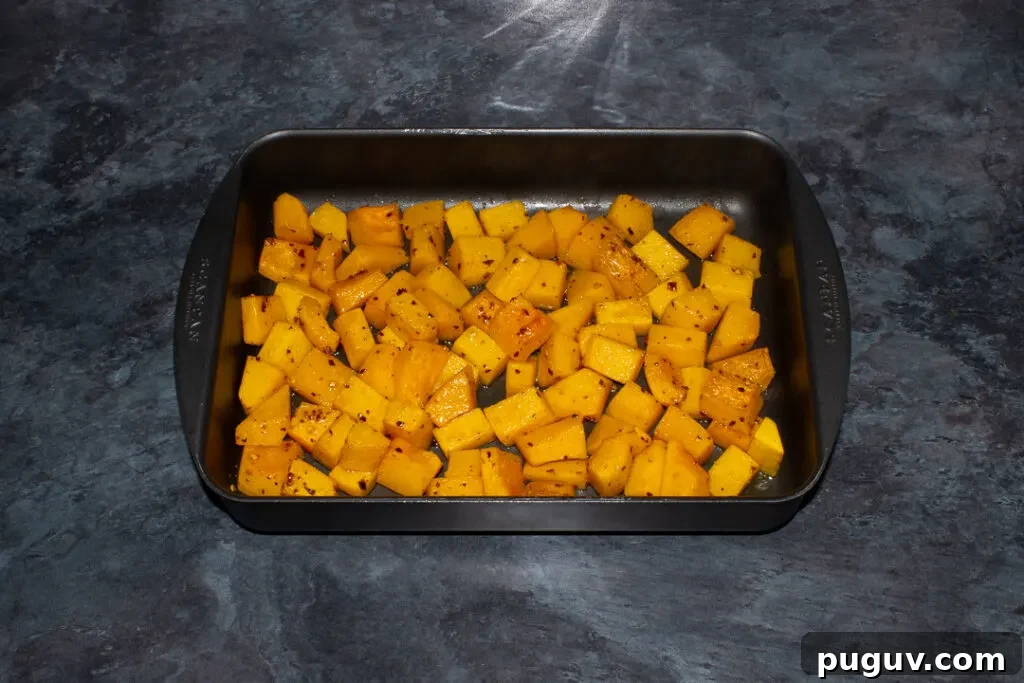 Roasted cubes of butternut squash that have been seasoned with chilli flakes in a roasting pan. Set on a kitchen worktop.