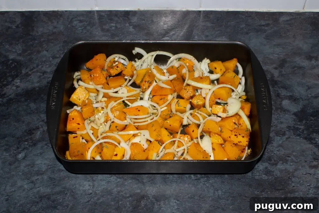 Cubes of butternut squash roasted with chilli flakes in a roasting dish. Sliced onion and garlic has been added to the pan. Set on a kitchen worktop.