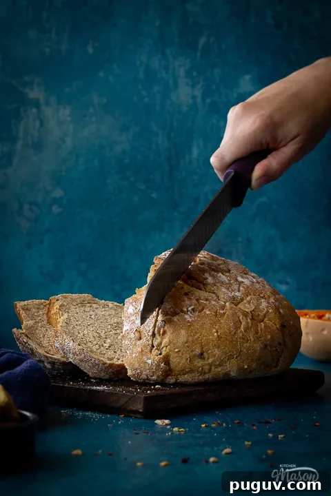 A hand reaching in to slice a loaf of sourdough bread with a bread knife on a dark wood chopping board. All set against a teal backdrop, there's also half a butternut squash, a pot of croutons, a deep blue linen napkin, chilli flakes and breadcrumbs in the background.