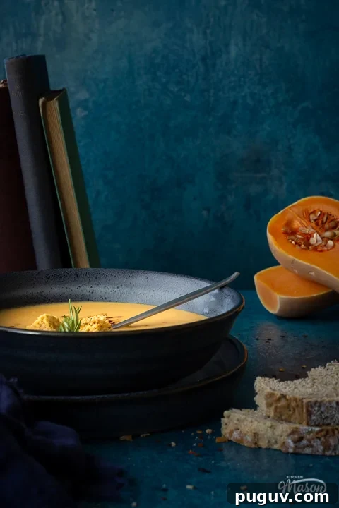 Front view of a bowl of spicy roast butternut squash soup with a spoon in it set over a plate. There is a dark blue linen napkin, some books stacked up at the back, two halves of a butternut squash and some bread in the background. Set on a teal backdrop.