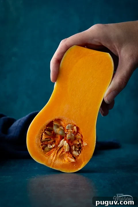 Close up front view of someone holding half a butternut squash against a teal backdrop. There is a dark blue linen napkin scrunched up in the background.