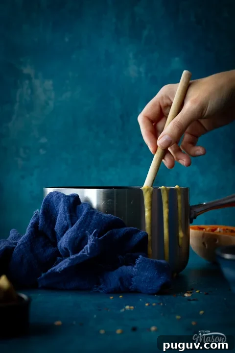 Someone stirring with a wooden spoon in a saucepan of butternut squash soup set against a teal backdrop. There are drips of soup down the side of the pan, half a butternut squash, a pot of croutons and a blue linen napkin in the backgtround.