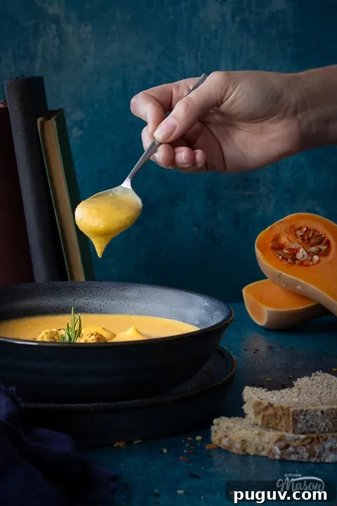 Front view of a bowl of spicy roast butternut squash soup with a hand holding up a spoon filled with soup above it set over a plate. There is a dark blue linen napkin, some books stacked up at the back, two halves of a butternut squash and some bread in the background. Set on a teal backdrop.