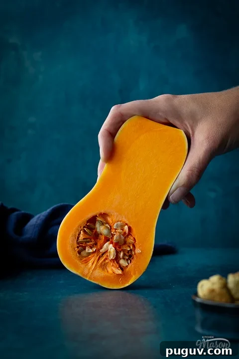 Distanced front view of someone holding half a butternut squash against a teal backdrop. There is a dark blue linen napkin scrunched up and a pot of croutons in the background.