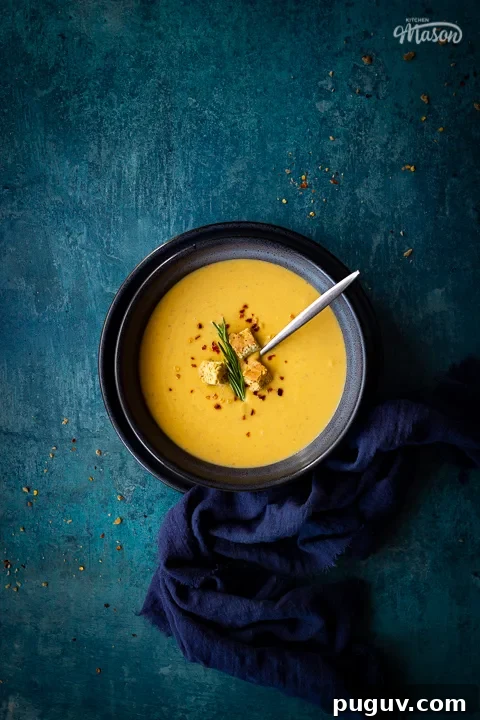 Flat lay view of spicy roast butternut squash soup with a spoon in a blue bowl set on a blue plate with a dark blue linen napkin on the side. Set on a teal backdrop, there are a few chilli flakes and breadcrumbs scattered in the background.