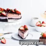 Slice of chocolate strawberry cheesecake on a blue plate in front of the rest of the cake, a bowl of strawberries, a ceramic pitcher and a glass of wine.