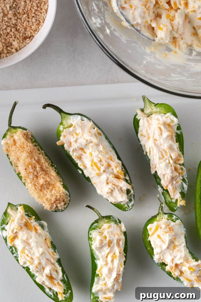 Crispy Air Fryer Jalapeño Poppers: Cream Cheese Stuffed Bites 5 Process of jalapeños being filled and breaded on a white cutting board. Panko bread crumb in a bowl on the upper left and cream cheese mixture on the upper right.
