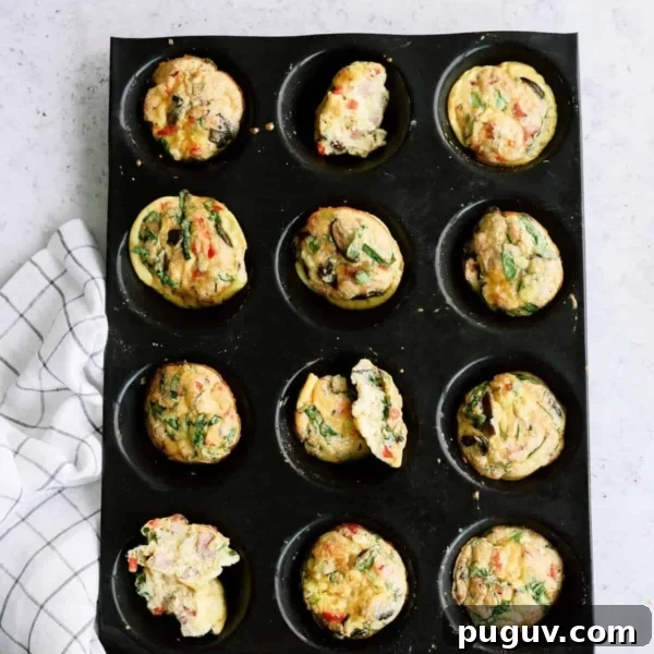 Muffin tray with twelve cooked egg muffins, some with spinach and vegetables, on a light surface next to a checkered cloth.