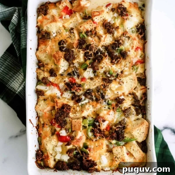 Baked casserole with bread cubes, bell peppers, onions, melted cheese, and browned sausage in a white rectangular dish on a green checkered cloth.