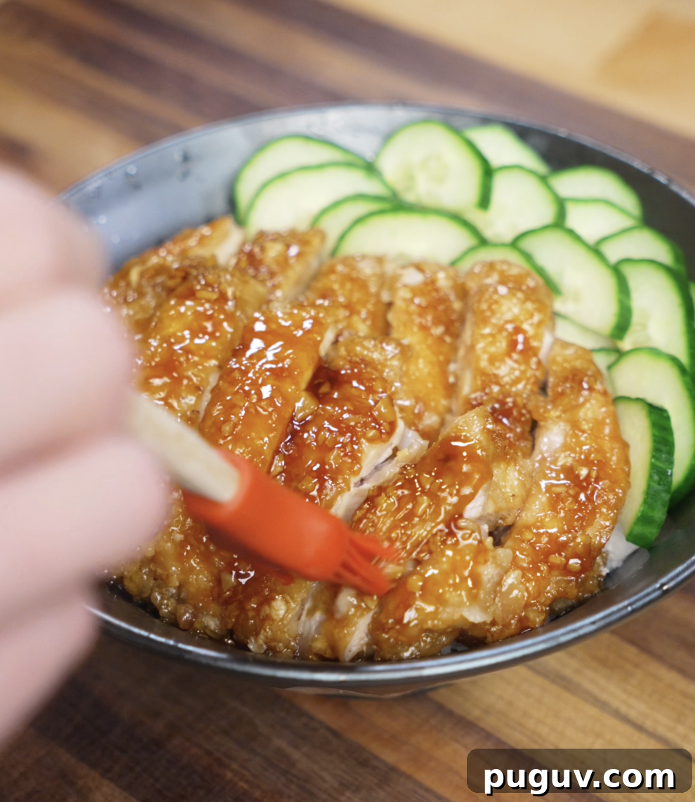 Crispy Honey Garlic Chicken Recipe — Sticky Glazed Chicken Drumsticks 8 A hand uses a red brush to apply extra honey garlic glaze over sliced crispy chicken thighs, adding shine and flavor to the final rice bowl topped with cucumbers.