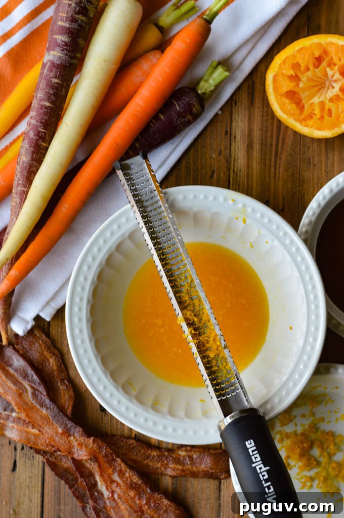 Raw rainbow carrots sitting next to a bowl of orange juice, bacon and maple syrup with a zester on top of the bowl.