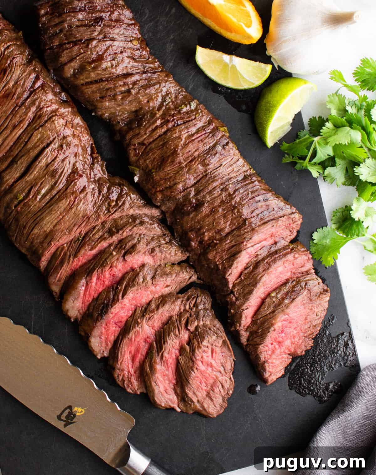 sliced carne asada made in the oven on a black cutting board with knife and citrus fruits photographed from above.