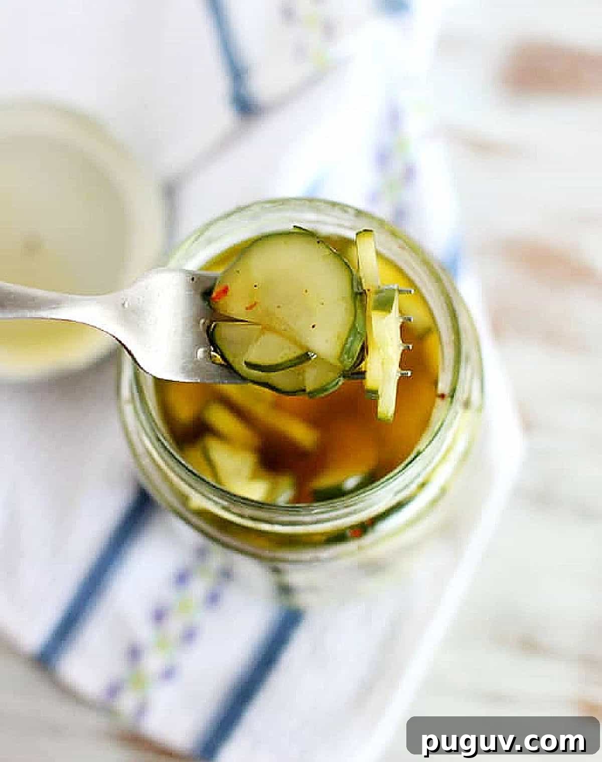 Quick Refrigerator Pickled Cucumbers: Tangy Crunch in 24 Hours 6 White and blue cloth beneath a jar with pickled cucumbers and a silver fork.