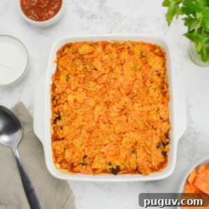 A white square baking dish filled filled with Doritos casserole is surrounded by small bowls filled with salsa, Greek yogurt and Doritos. A serving spoon and brown napkin sit to the side.