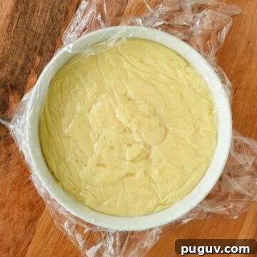 Thickened pastry cream in a white bowl covered with plastic wrap.