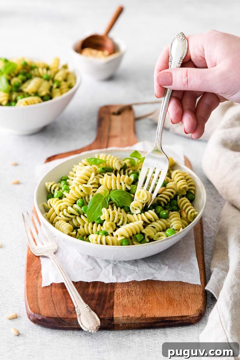 Creamy Pea Pasta with Garlic and Parmesan 3 Woman's hand holding a fork in bowl of rotini pesto pasta with veggies, sitting on top of a wooden serving board.