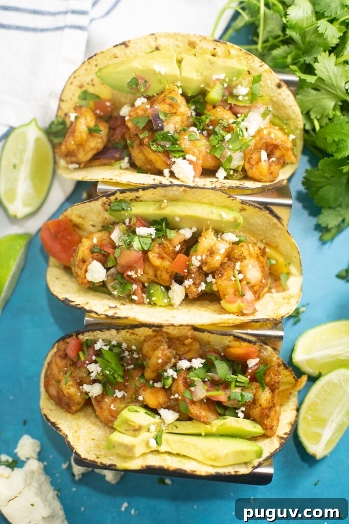 Sheet Pan Shrimp Fajitas Recipe for Quick Weeknight Dinners 2 Overhead view of three sheet pan shrimp fajitas in corn tortillas topped with fresh cilantro, queso fresco, and avocado slices, and surrounded by lime wedges, a bunch of fresh cilantro, and a linen napkin.