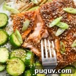 30-Minute Garlic-Ginger Glazed Salmon Recipe (Gluten-Free) 15 Caramelized garlic ginger salmon filet on white rice with a side of crunchy sesame cucumber salad, on a wide ceramic plate, with a fork digging into the salmon to show the tender interior.