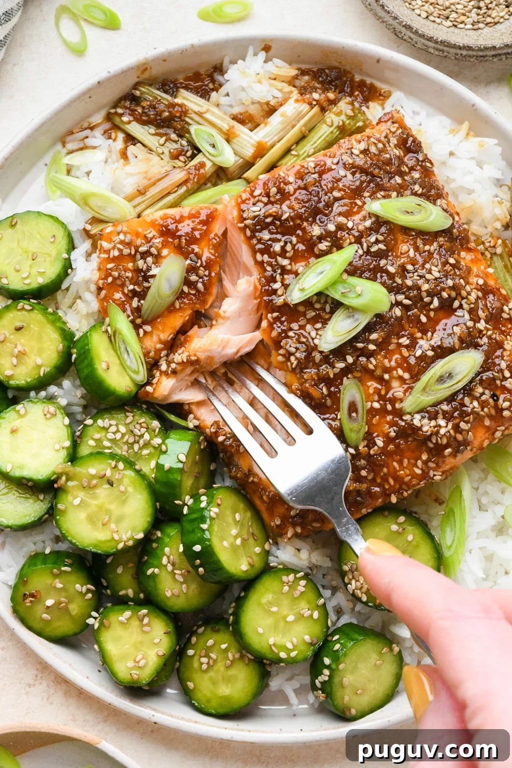 30-Minute Garlic-Ginger Glazed Salmon Recipe (Gluten-Free) 3 Caramelized garlic ginger salmon filet on white rice with a side of crunchy sesame cucumber salad, on a wide ceramic plate, with a fork digging into the salmon to show the tender interior.