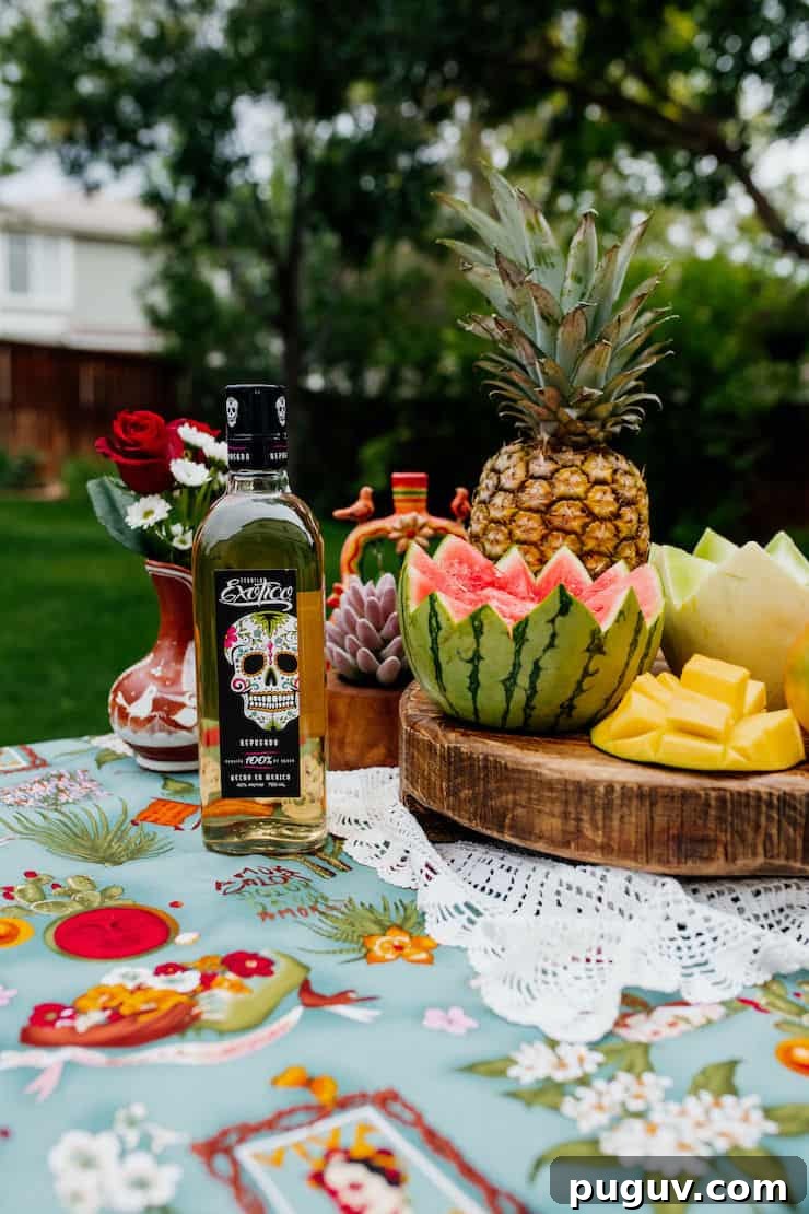 Frida Kahlo-Inspired Sangria Recipe for Parties 5 exotic tequila bottle with melons and a pineapple on a wooden pedestal on a white doily and teal tablecloth outside